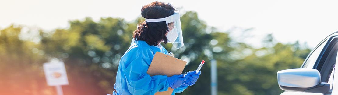 A woman stands in PPE ready to give a COVID test to an upcoming vehicle