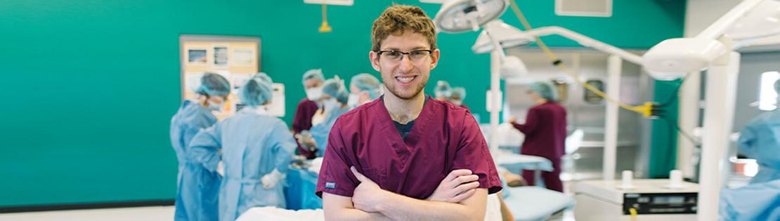 一名外科技术学生在皮马外科课上摆姿势拍照，同学们在他身后练习